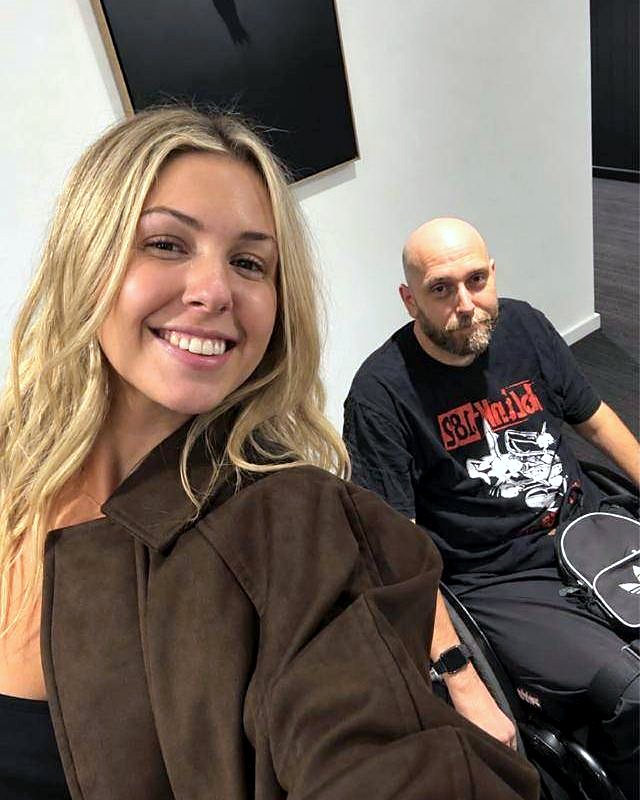 Abigail taking a selfie with a smiling man who uses a wheelchair, at an art gallery.
