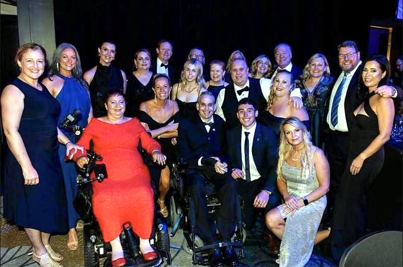 A large group in formal wear, including two people in power wheelchairs, smiling together at an awards night.