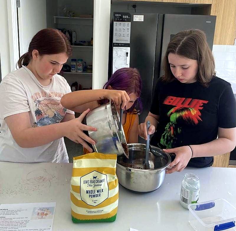 Two young women baking together in a kitchen, one pouring from a bowl while the other stirs.