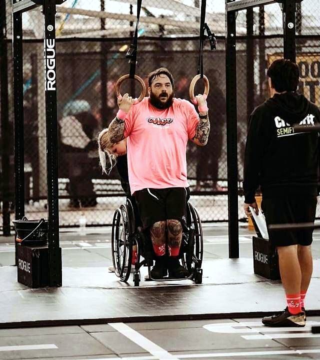 An adaptive CrossFit athlete in a pink shirt doing ring pull-ups from his wheelchair at a competition.