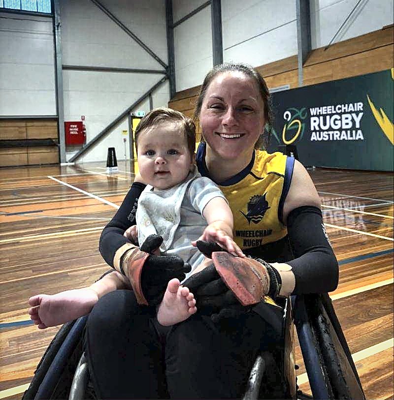 A smiling wheelchair rugby player in a yellow jersey holding a baby on her lap at a Wheelchair Rugby Australia court.