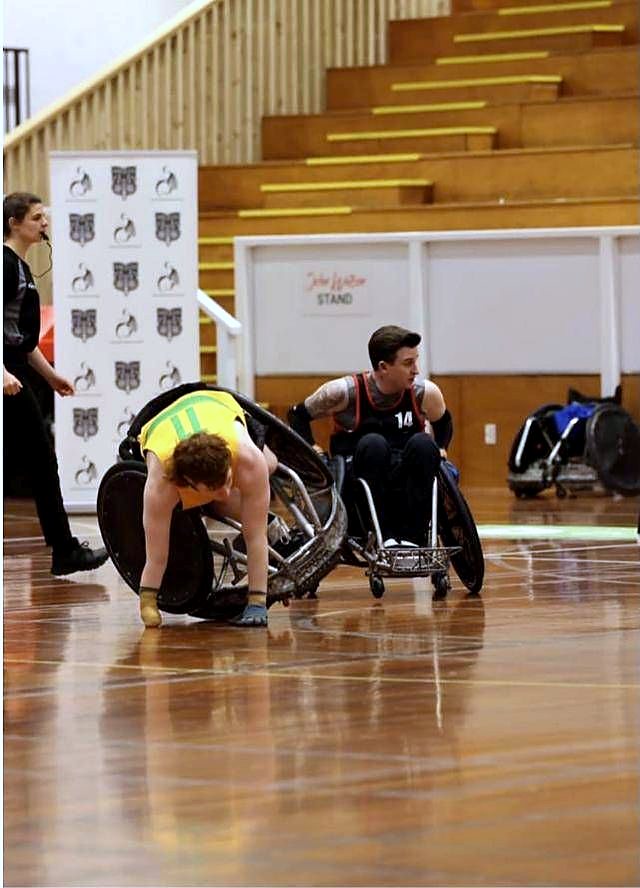 A wheelchair rugby player in a green and gold jersey leaning low out of his chair to reach the court during a game.